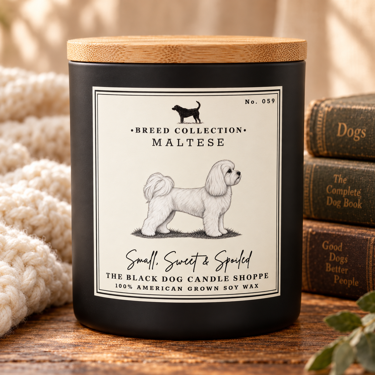 A black candle jar with a light wood lid sits on a wooden surface, wrapped in a cream knit blanket on the left and next to stacked dog books on the right. The front label reads 'Breed Collection - Maltese' and 'Small, Sweet & Spoiled' from 'The Black Dog Candle Shoppe'.