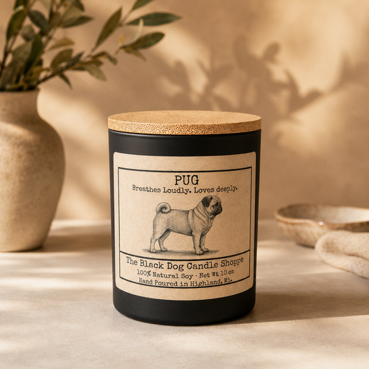Matte black soy candle with vintage-style pug illustration label and bamboo lid, displayed in a warm minimal setting with soft neutral tones and natural light.