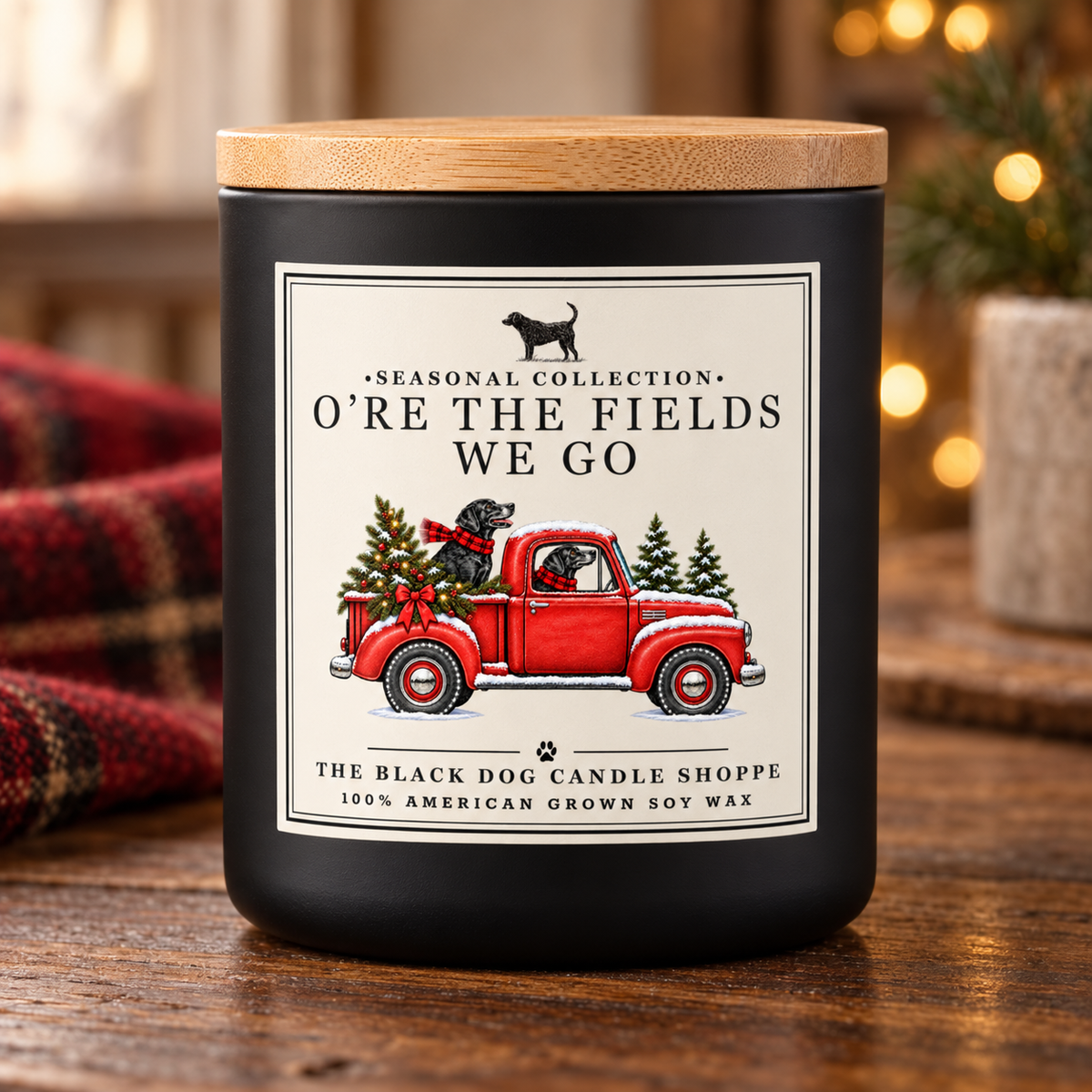 A black candle jar with a light wood lid sits on a wooden table. The front label reads “Seasonal Collection” and “O’re the Fields We Go,” with an illustration of a red pickup truck carrying Christmas trees.