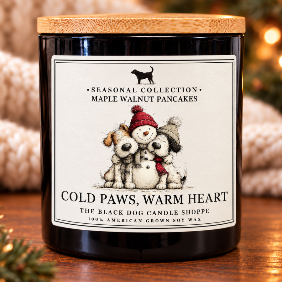 Black candle with a bamboo lid and a “Cold Paws, Warm Heart” label featuring two dogs beside a snowman. The Maple Walnut Pancakes candle sits on a wooden table against a cozy holiday backdrop.