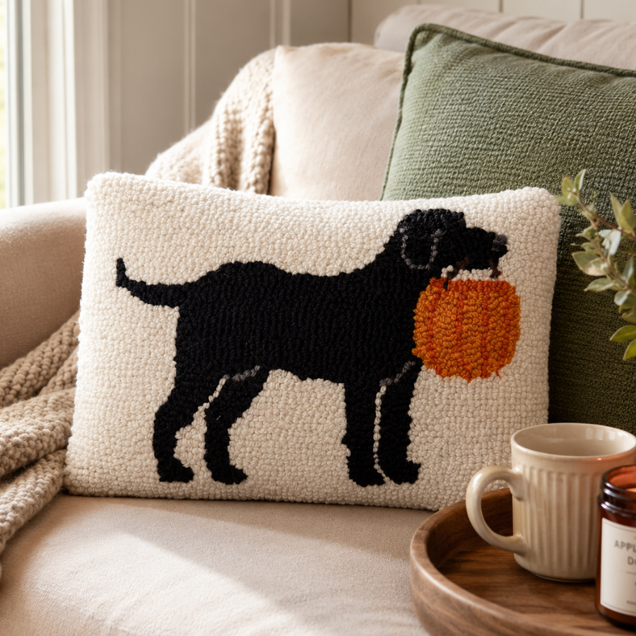 A textured throw pillow featuring a black dog holding an orange pumpkin, styled on a beige sofa next to a green pillow and a wooden tray with a mug.