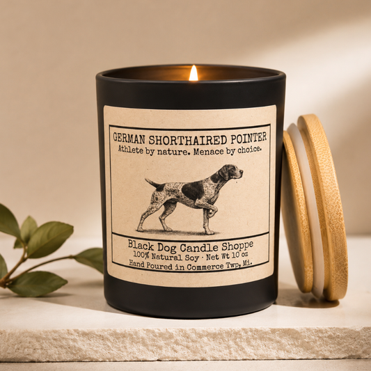 Minimal cozy product photo of a lit German Shorthaired Pointer soy candle in a matte black vessel with bamboo lid beside it, styled on a light stone surface with soft neutral lighting and subtle greenery against a warm beige background.