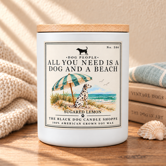 A white soy wax candle jar with a light wood lid sits on a wooden surface, surrounded by a cozy styled setting including a cream knit blanket, a stack of vintage dog books, a seashell, and a folded blue-green cloth. The front label features a beach illustration with a Dalmatian, an umbrella, and the text 'ALL YOU NEED IS A DOG AND A BEACH' and 'SUGARED LEMON'.