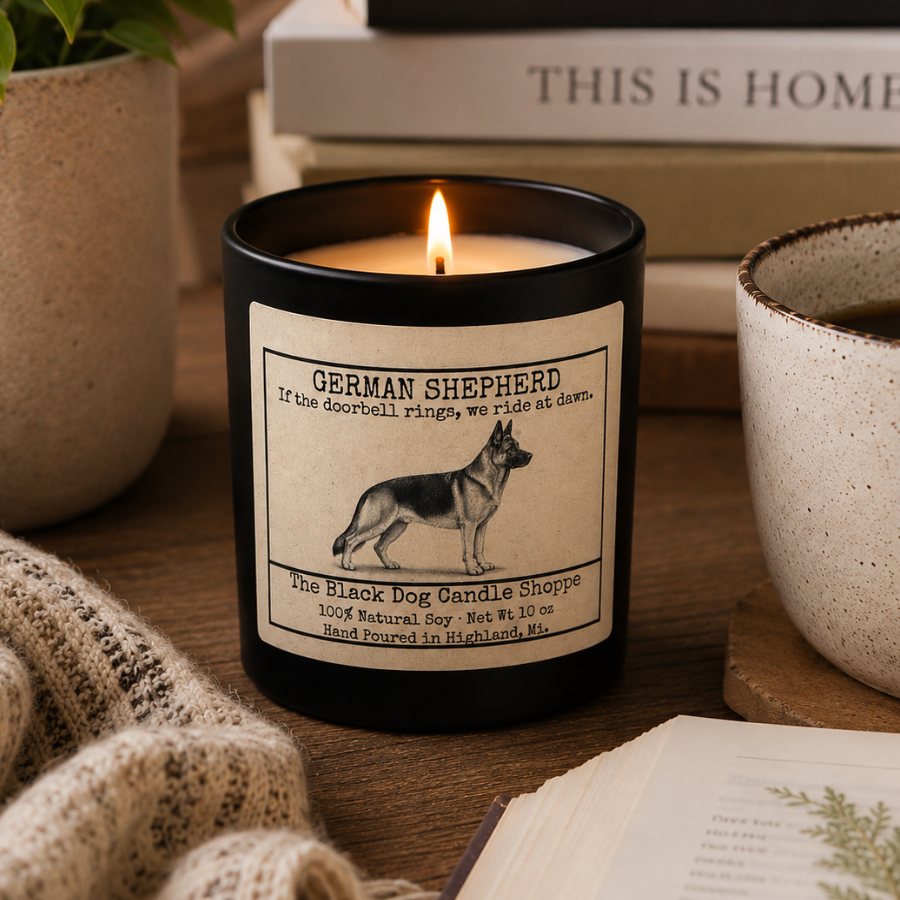 Matte black German Shepherd soy candle with vintage-style dog illustration label and bamboo lid, displayed in a warm cozy lifestyle setting with books, coffee, and soft neutral decor.