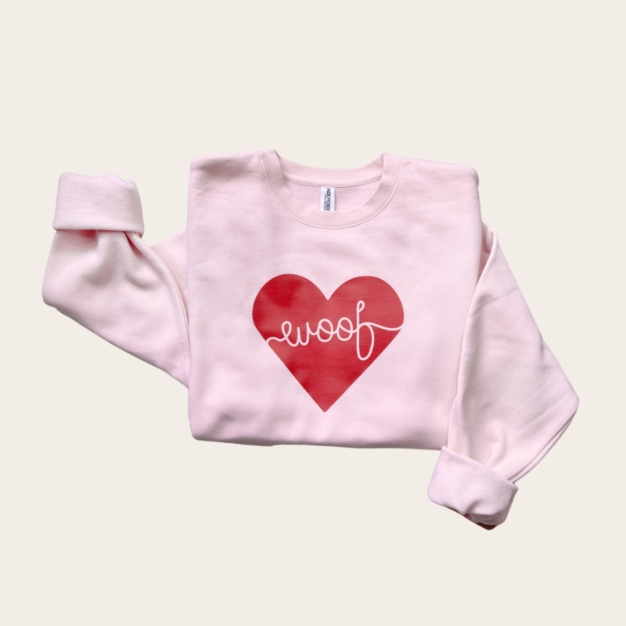 Light pink crewneck sweatshirt with red heart graphic and “woof” script, cozy dog lover apparel laid flat on neutral background